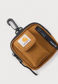 Μικρό καφέ υφασμάτινο πουγκί με μαύρο φερμουάρ, μικρή μπροστινή τσέπη με το λογότυπο της Carhartt και μαύρο κλιπ για να το κρεμάτε.