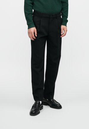 Zwarte op maat gemaakte broeken met een rechte pasvorm, een gladde textuur, voorzien van riemlussen en een enkele knoopsluiting. Draagt met zwarte nette schoenen.