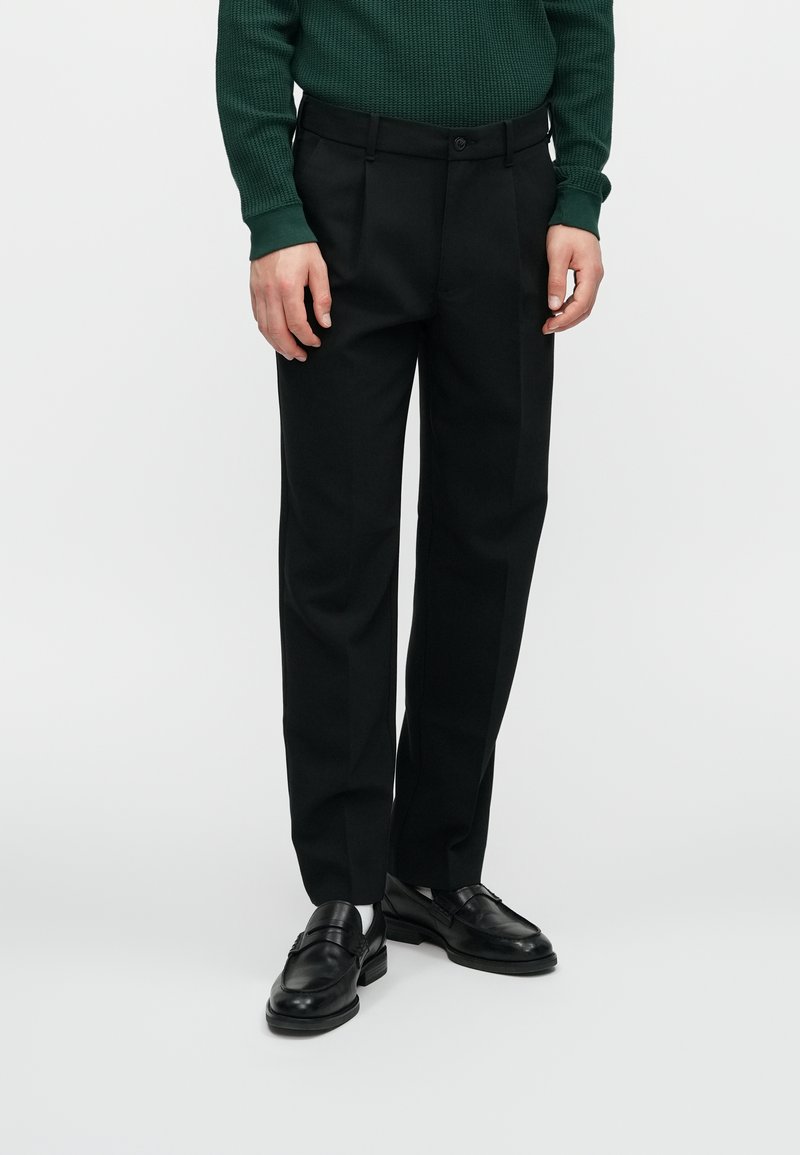 Zwarte op maat gemaakte broeken met een rechte pasvorm, een gladde textuur, voorzien van riemlussen en een enkele knoopsluiting. Draagt met zwarte nette schoenen.