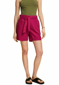 Bermudas de lino magenta con cinturón, que presentan un corte relajado y bolsillos laterales, combinadas con una blusa verde ajustada y sandalias negras.