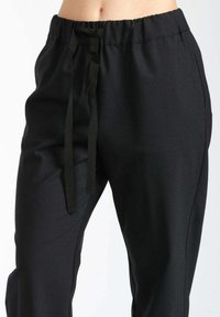 Pantaloni neri con elastico in vita e lunghi lacci, aderenti in vita e morbidi sulle gambe, mostrati sulla parte bassa del torso di una persona.