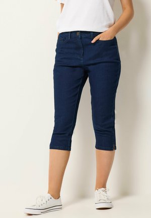 Kvinde iført mørkeblå cropped skinny jeans med små sideslids, hvide sneakers og en hvid t-shirt med korte ærmer, hånd i lommen.