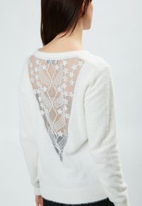 Maglione bianco lavorato a maglia con una morbida e pelosa texture e un pannello in pizzo triangolare sul retro con motivi a stella, creando un design a strati.