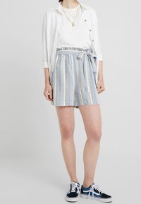 Blå och vita randiga shorts med slips, kombinerat med en vit skjorta, lager av halsband och marinblå sneakers.