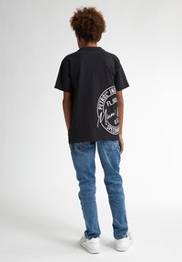 Sort bomulds T-shirt med et rundt hvidt logo på bagsiden, parret med blå jeans og hvide sneakers. Korte ærmer, afslappet pasform.