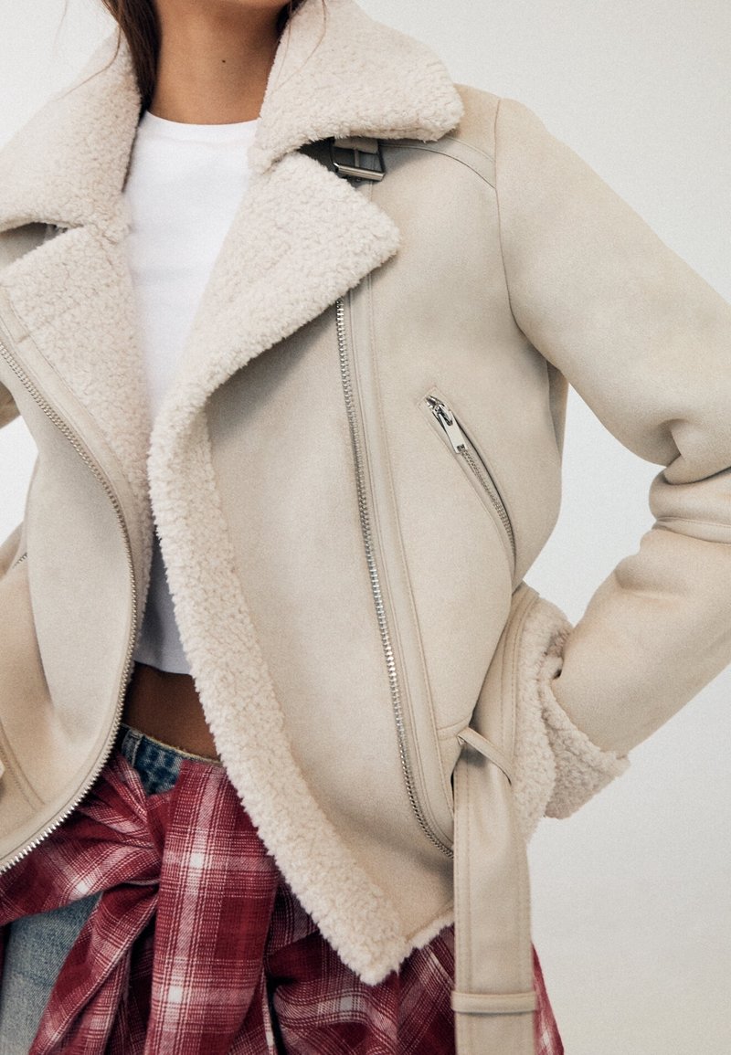 Veste en shearling beige avec fermetures éclair argentées sur une chemise blanche, chemise à carreaux rouge nouée autour de la taille, personne posant avec la main sur la hanche.