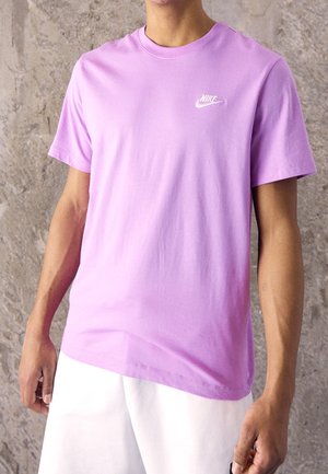 Mężczyzna w jednokolorowej lawendowej koszulce Nike i białych szortach stoi na tle teksturowanej szarej ściany.