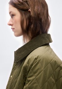 Νεαρή γυναίκα με καστανά μαλλιά που φοράει ένα λαδί καπιτονέ μπουφάν με γιακά από σκούρο πράσινο κορδούρα, στραμμένη προς τα αριστερά σε απλό φόντο.