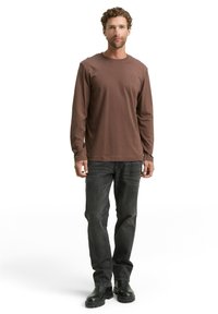 Bruine katoenen overhemd met lange mouwen en een ronde halslijn, gecombineerd met donkergrijze denim jeans en zwarte laarzen, staand tegen een witte achtergrond.