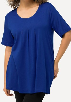 Curly-haired person wearing a loose-fitting blue short-sleeve top with front pleats, paired with black bottoms, standing with one hand on hip.