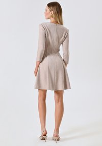 Robe beige à manches trois-quarts avec un corsage ajusté et une jupe évasée. Dotée d'une fermeture éclair au dos et d'une texture lisse. Accompagnée de sandales à talons.