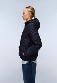 Marinblå vadderad jacka med huva, matlad struktur, framficka och elastiska ärmslut. Bärs med en randig skjorta och raka jeans.