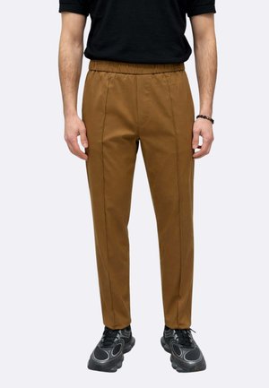 Uomo che indossa pantaloni marroni a vita elastica con dettagli a cucitura, scarpe da ginnastica nere e una maglietta nera a maniche corte, in piedi contro uno sfondo semplice.