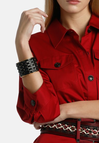 Red button-up shirt with two chest pockets, cinched sleeves, and a black woven cuff bracelet with gold accents. Textured belt at waist.
