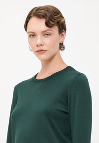 Pull en maille long à manches longues de couleur vert foncé avec un col rond. Texture lisse et détail de couture subtil le long du col.