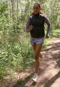 Un débardeur noir sans manches sur un haut à manches longues gris, associé à un short bleu clair et des chaussures de course beigées, le tout dans une forêt verte.