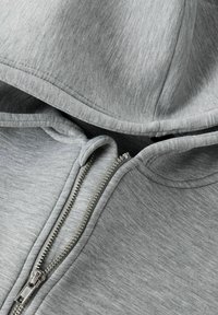 Pelēks zip-up kapucis ar mīkstu heather audumu, kapuci un sudraba rāvējslēdzēju pie priekšējā atvēruma.
