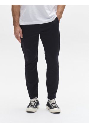Man draagt zwarte taps toelopende broek, zwart-witte sneakers en een wit T-shirt, staand met één hand in de zak.