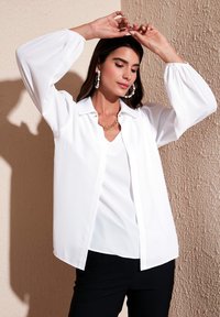 White long-sleeve blouse with a button-up front, collared neckline, and balloon sleeves, layered over a white v-neck top. Black pants visible.