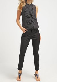 Mujer con una blusa negra sin mangas con volantes, pantalones negros ajustados y sandalias de tacón azul marino con correas en el tobillo, de pie con una mano en el bolsillo.