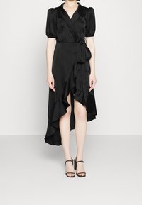 Robe portefeuille en satin noir avec manches courtes bouffantes, ourlet asymétrique et détails à volants. Portée avec des talons noirs à lanières.