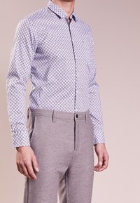 Man som bär en vit mönstrad skjorta med långa ärmar instoppad i ljusgrå byxor mot en neutral bakgrund.