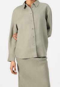 Chemise gris clair à manches longues avec boutons, en lin, avec un col, une texture lisse et un ourlet légèrement arrondi. Portée avec une jupe assortie.