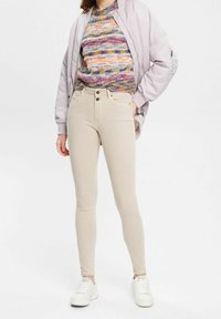 Pantalones ajustados beige con bolsillos delanteros, cierre de botón y trabillas para cinturón, combinados con un suéter de punto texturizado multicolor y una chaqueta bomber ligera.