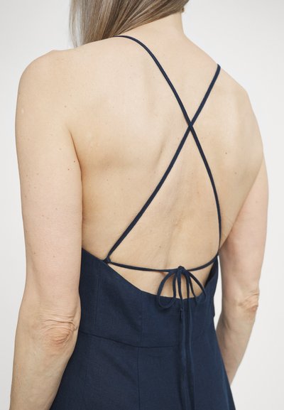 Robe dos nu bleu marine avec de fines bretelles croisées et un détail noué à la taille, dotée d'une texture lisse et d'un design minimaliste.