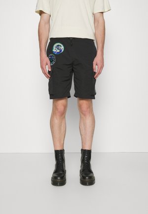 Bermuda cargo noirs en tissu léger, avec des patchs circulaires colorés et une taille ajustable à cordon. Portés avec des bottes noires.