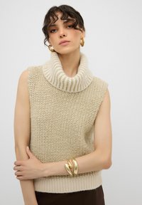 Maglione smanicato a collo alto beige lavorato a maglia con colletto a coste, caratterizzato da un motivo testurizzato e un orlo aderente. Modella che indossa bracciali dorati.