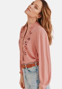 Helles rosa Blusenshirt mit langen, gepufften Ärmeln, das eine florale Stickerei entlang der Vorderseite und einen knöpfbaren Ausschnitt hat. Kombiniert mit Jeans.