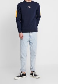 Uomo che indossa una felpa blu navy con accenti gialli e bianchi sulle maniche, jeans azzurri arrotolati e scarpe da ginnastica nere a bassa cima con lacci bianchi.