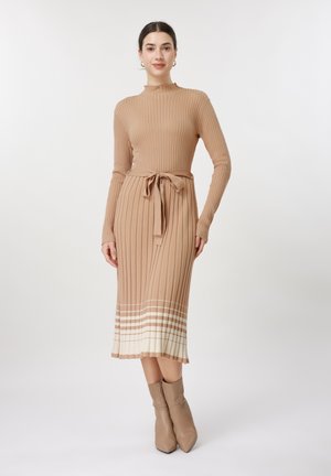 Žena stojaca, oblečená v béžových rebrovaných pletených šatách s bielymi pruhmi na leme a v odpovedajúcich členkových čižmách, so zviazaným opaskom v páse.