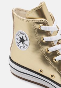 Zlatne metalik Converse Chuck Taylor visoke tenisice s bijelim vezicama, crnom zvjezdicom kao logom, i gumenim đonom sa smešnim dno.