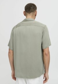 Camisa de manga corta de tela verde apagada, textura suave, cuello abierto y dobladillo recto. La parte trasera presenta un diseño simple sin adornos.