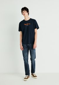 Jūros mėlynos spalvos medvilninis marškinėliai su oranžiniu "Pepe Jeans" logotipu, derinami su tamsiai mėlynomis džinsais ir rudais zomšiniais sportbačiais ant vienspalvio fono.