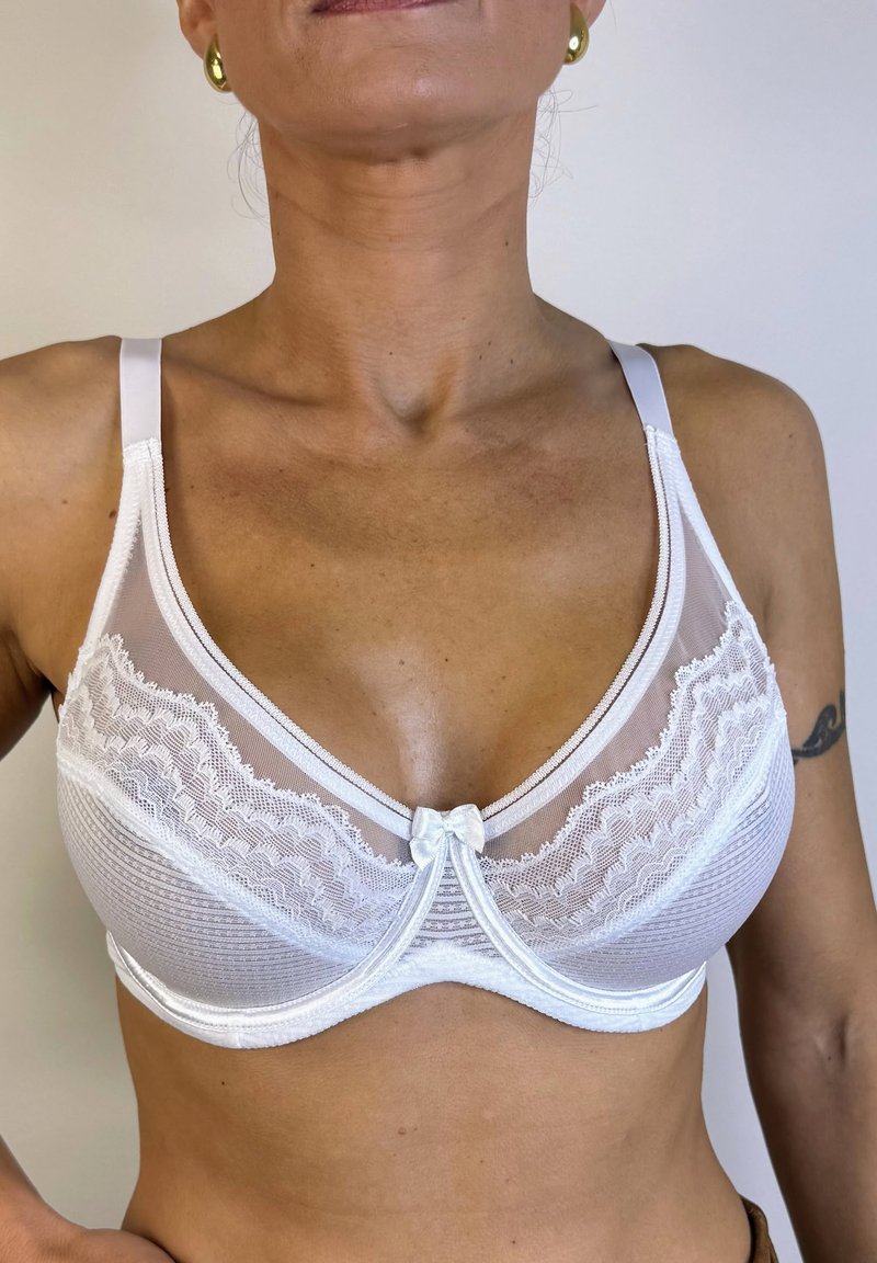 Sostén de encaje blanco con un panel superior transparente, detalles de encaje con forma de ondas, un lazo central decorativo y tirantes ajustables.