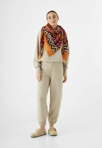 Vrouw staat in beige loungewear, beige instappers en een groot kleurrijk geometric patroon sjaal in oranje, roze en bruine tinten.