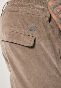 Bruine corduroy broek met een achterzak en een metalen logo tab. De stof heeft een zachte, gestructureerde uitstraling en verticale lijnen.