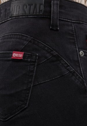 Detaljan prikaz stražnjeg džepa crnih traperica s crvenom tkanom oznakom "BIG STAR" i crnim kožnim logom na pojasu.