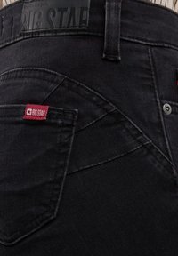 Detaljan prikaz stražnjeg džepa crnih traperica s crvenom tkanom oznakom "BIG STAR" i crnim kožnim logom na pojasu.