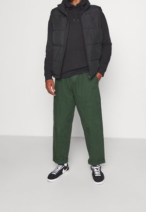 Czarna pikowana kamizelka na czarnej bluzie z kapturem, zestawiona z luźnymi zielonymi spodniami typu cargo oraz czarnymi sneakersami z białymi akcentami. Prosty i funkcjonalny design.