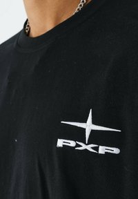 Camiseta de algodón negra con un logo bordado en blanco "PXP" y un diseño de estrella en el lado izquierdo del pecho. Mangas cortas y cuello redondo.
