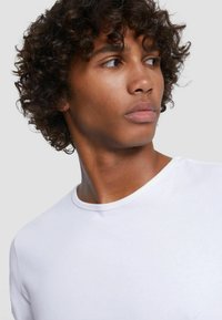 White short-sleeve t-shirt featuring a crew neck and smooth fabric. Close-up of a model with curly hair, side profile against a light background.