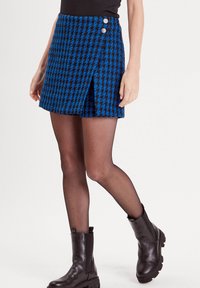 Mini-jupe en vichy bleu et noir avec un design enroulé, dotée de deux boutons gris et d'une fente latérale. Associée à des bottines noires et des collants.