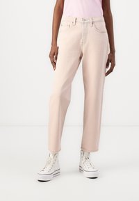 Una persona vistiendo jeans rectos de color rosa claro, una camiseta de rosa pálido y zapatillas de plataforma altas blancas frente a un fondo blanco.