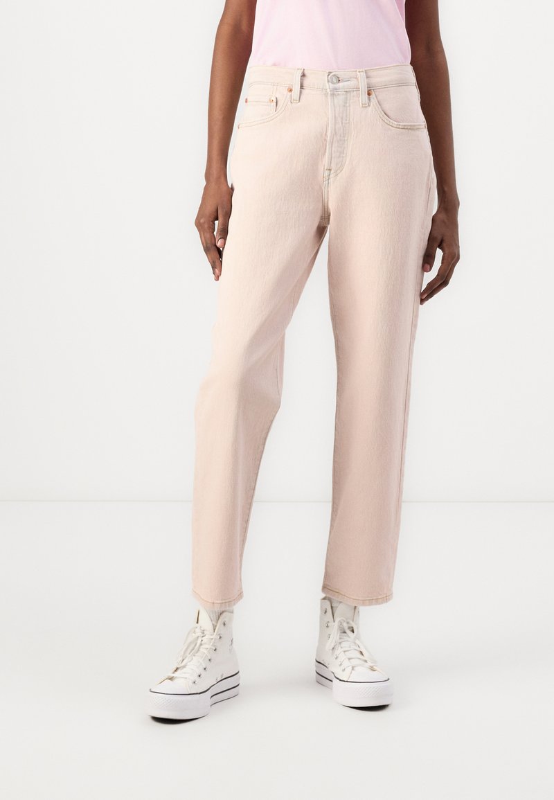 Una persona vistiendo jeans rectos de color rosa claro, una camiseta de rosa pálido y zapatillas de plataforma altas blancas frente a un fondo blanco.