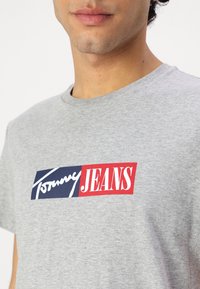 Pilkos spalvos medvilninė marškinėlė su stačiakampiu logotipu tamsiai mėlynos ir raudonos spalvų su žodžiais "Tommy" ir "JEANS" dėl skirtingų šriftų. Apvalus iškirpimas.