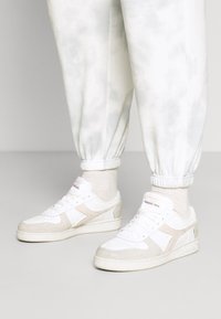 Bílé tenisky s texturovaným béžovým akcentem, které mají perforovanou špičku, tkaničky a logo na boku. Noseny s lehkými, vzorovanými tepláky.